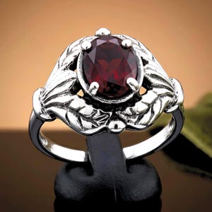 May include: A silver ring featuring an oval-cut deep red gemstone set in the center. The band and setting are crafted with a detailed relief pattern of stylized leaves that surround the stone, held in place by four small metal prongs.