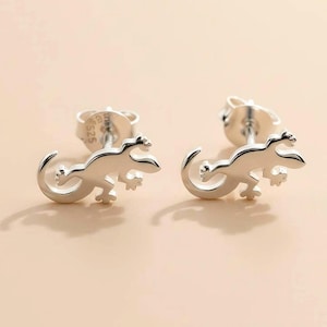 May include: A pair of silver stud earrings in the shape of a lizard. The earrings are detailed with a curved tail and four legs. The earrings are on a light pink background.