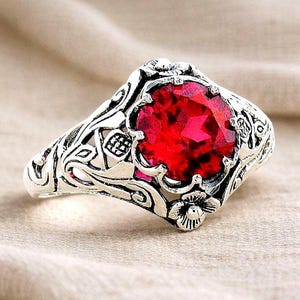 May include: A silver ring with a large, faceted, red gemstone. The ring features an ornate floral and scrollwork design around the gemstone and on the band. The ring is set against a neutral background.