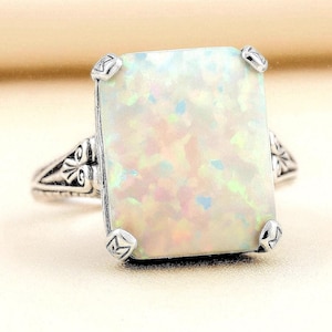 May include: A rectangular white opal ring set in an oxidized silver band. The stone features iridescent flashes of pink, green, and blue. The silver band displays intricate engraved detailing on the sides and is secured by four corner prongs.