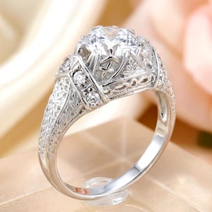 May include: A silver-toned ring featuring a large, round, clear center stone held in a prong setting. The band and gallery are decorated with intricate, engraved leaf-like patterns and small, round, clear accent stones along the shoulders.