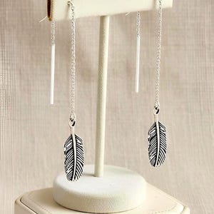 May include: Silver feather earrings with long chain and bar accents. The earrings are displayed on a white jewelry stand. The feather pendants have detailed black and white feather patterns. The earrings are a delicate and stylish accessory.