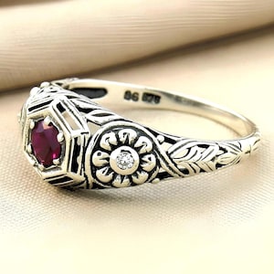 May include: A silver ring featuring a central hexagonal setting with a round red gemstone, flanked by a circular floral motif containing a small clear stone. The band displays intricate engraved leaf patterns and the text 925 878 inside.