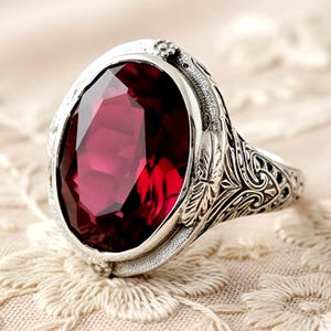 May include: A silver ring featuring an oval, faceted, deep red gemstone. The ring has an ornate, engraved design on the band and around the gemstone setting, with floral and leaf motifs. The ring is set against a cream-colored, lace-like background.