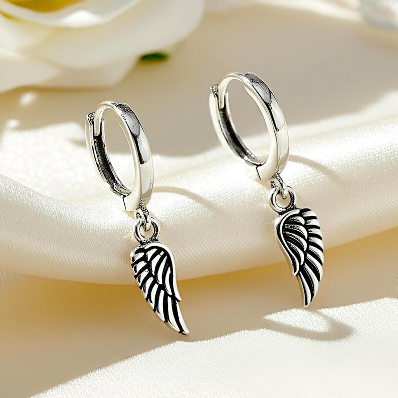 May include: A pair of silver hoop earrings with dangling wing charms. The earrings have a polished finish and a hinged closure. The wing charms are detailed with etched lines, creating a feather-like appearance. The earrings are displayed on a white satin surface.