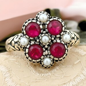 May include: A silver ring featuring a floral design with five round, deep red gemstones and six smaller, white pearls. The ring's band has an ornate, swirling pattern. The gemstones are set in a detailed, silver setting.
