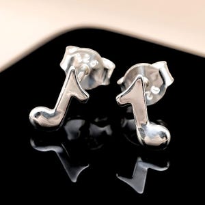 May include: A pair of silver-colored musical note stud earrings. Each earring is shaped like a musical eighth note, with a curved lower portion and a straight stem. The earrings have a shiny, reflective surface.