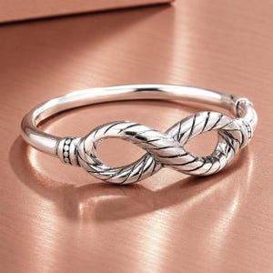 May include: A silver-colored bangle bracelet with an infinity knot design. The bracelet features a textured rope-like pattern on the infinity symbol and a smooth, polished finish on the band. The bracelet is displayed on a copper-colored surface.