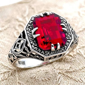 May include: A sterling silver ring with an octagonal, bright red gemstone. The ring features an ornate, detailed design around the gemstone and on the band. The ring is set on a cream-colored, textured surface.