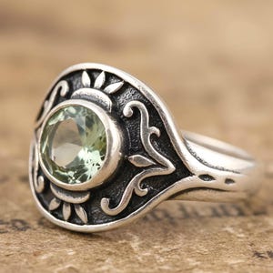 May include: A silver ring with a round, light green gemstone set in a decorative, dark-toned bezel. The ring features an ornate design with floral and sunburst-like elements. The band is smooth and polished.