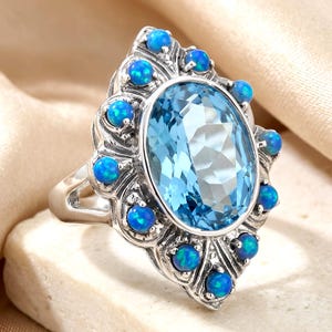 May include: A silver ring featuring a large, oval-cut, light blue gemstone at the center, surrounded by a halo of twelve small, round, iridescent blue cabochon stones set in a decorative, scalloped metal frame with a polished finish.