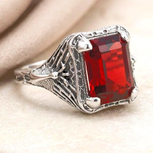 May include: A silver-toned ring featuring a large, rectangular, faceted red gemstone set in a prong setting. The band and setting display an intricate, textured, web-like metal design with darkened recessed areas that highlight the detailed pattern.