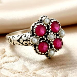 May include: A silver ring with a floral design, featuring five round, bright red gemstones and six small, white pearls. The ring's band has an ornate, swirling pattern.