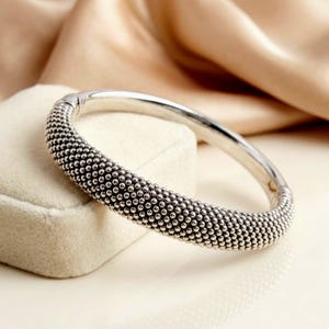 May include: A silver bangle bracelet with a textured beaded design. The bracelet is round and has a series of small, evenly spaced beads covering the majority of the band. The bracelet is displayed on a white surface.
