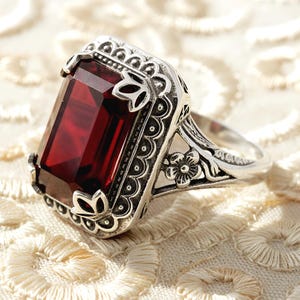 May include: A silver ring with a large, rectangular, deep red gemstone. The ring has an ornate, filigree design with floral and scroll details around the stone and on the band. The setting is detailed with small, decorative elements.