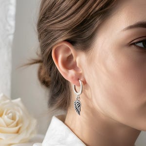 May include: A silver hoop earring with a small, dangling wing charm. The wing is detailed with black lines, creating a feather-like appearance. The earring is worn on the ear of a person wearing a white shirt.