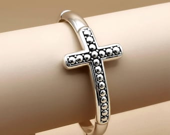 May include: A silver-toned cross-shaped ring with a beaded design. The cross is centered on the ring's band, with the beaded pattern outlining the cross shape. The ring is displayed against a neutral background.
