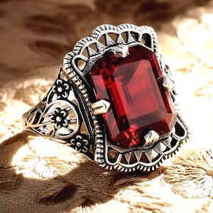 May include: A silver-toned ring featuring a large, rectangular, deep red gemstone. The ring has an ornate, filigree design with floral and geometric patterns. The gemstone is set in a decorative bezel, and the ring's band has intricate detailing.