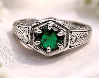 May include: A silver ring with an emerald-green gemstone in a hexagonal setting. The ring features intricate engraved patterns along the band, with a detailed design on the sides. The gemstone is the focal point, set with small prongs.