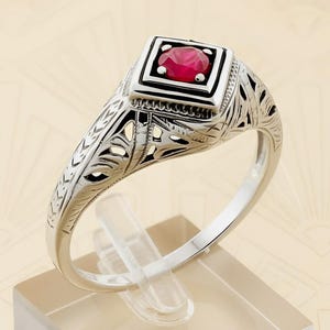 May include: A silver ring with a square, red gemstone. The ring has an ornate, filigree design on the band and around the gemstone setting. The ring is displayed on a clear stand.