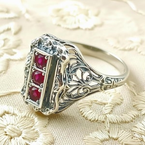 May include: A silver ring with a rectangular face featuring three red gemstones. The ring has an ornate, filigree design on the band and around the gemstone setting. The ring is set against a cream-colored, textured background.