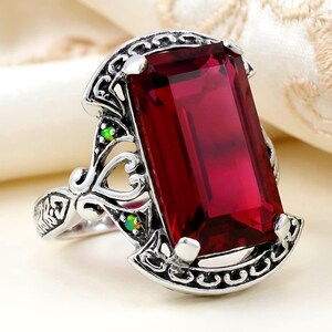 May include: A silver-toned ring featuring a large, rectangular, deep red gemstone. The ring has an ornate, filigree design with small green accent stones. The setting is detailed with scrollwork and decorative elements, creating a vintage or antique aesthetic.