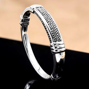 May include: A silver bangle bracelet with a textured design. The bracelet features a central band with a dotted pattern, framed by smooth, polished silver. The ends of the bracelet have decorative bands. The bracelet is displayed on a black surface.