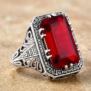 May include: A silver ring with a large, rectangular, red gemstone. The ring has an ornate, filigree design on the band and around the gemstone. The gemstone is a vibrant, deep red color.