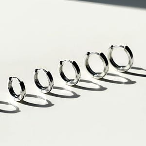 May include: Five silver hoop earrings of varying sizes are arranged diagonally on a white surface. The earrings are shiny and reflective, with a simple, minimalist design. The light creates shadows that emphasize their round shape.