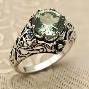 May include: A sterling silver ring featuring a large, round, faceted light green gemstone set in a prong setting. The band is crafted with intricate, raised floral and vine motifs that wrap around the sides, showcasing oxidized detailing.