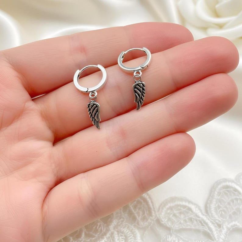May include: A pair of silver hoop earrings with a dangling wing charm. The earrings are shown on a hand, with a soft, white background. The wing charms have detailed feather designs.