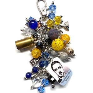May include: A silver keychain charm with various beads in blue, yellow, and clear. Charms include a brass bullet casing, a small trophy, and a tag with a portrait and text that reads "The murderer is right in this room."