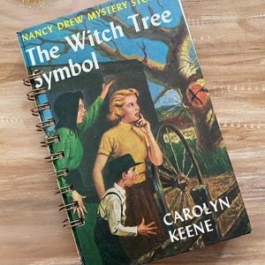 May include: Spiral-bound notebook featuring the cover of "The Witch Tree Symbol" by Carolyn Keene. The cover art depicts three people in front of a tree and a wagon wheel. The book has a blue and yellow color scheme.