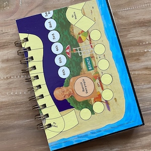 May include: Spiral-bound notebook with a colorful board game design. The game board features a beach scene with a blue ocean, yellow sand, and a cartoon character named Harold Dalrymple. The notebook has a wooden background.