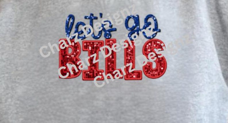 May include: A gray fabric with the text "let's go BILLS" embroidered in blue and red glitter. The text is in a playful, cursive font. The design is centered on the fabric.