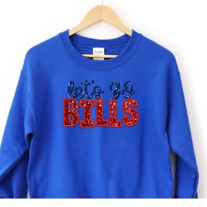May include: A royal blue sweatshirt with the text "let's go BILLS" in a glittery design. The text "let's go" is in blue glitter, and "BILLS" is in red glitter. The sweatshirt is hanging on a wooden hanger.