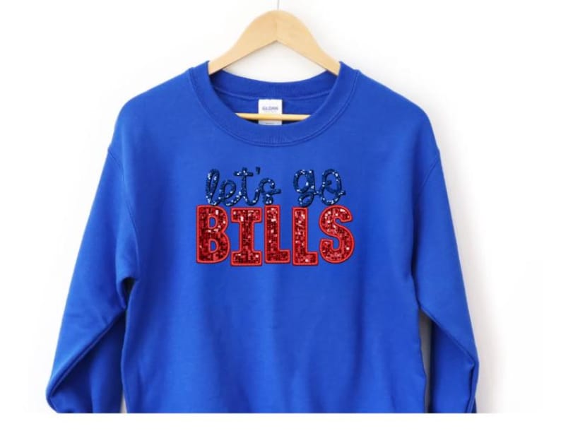 May include: A royal blue sweatshirt with the text "let's go BILLS" in a glittery design. The text "let's go" is in blue glitter, and "BILLS" is in red glitter. The sweatshirt is hanging on a wooden hanger.