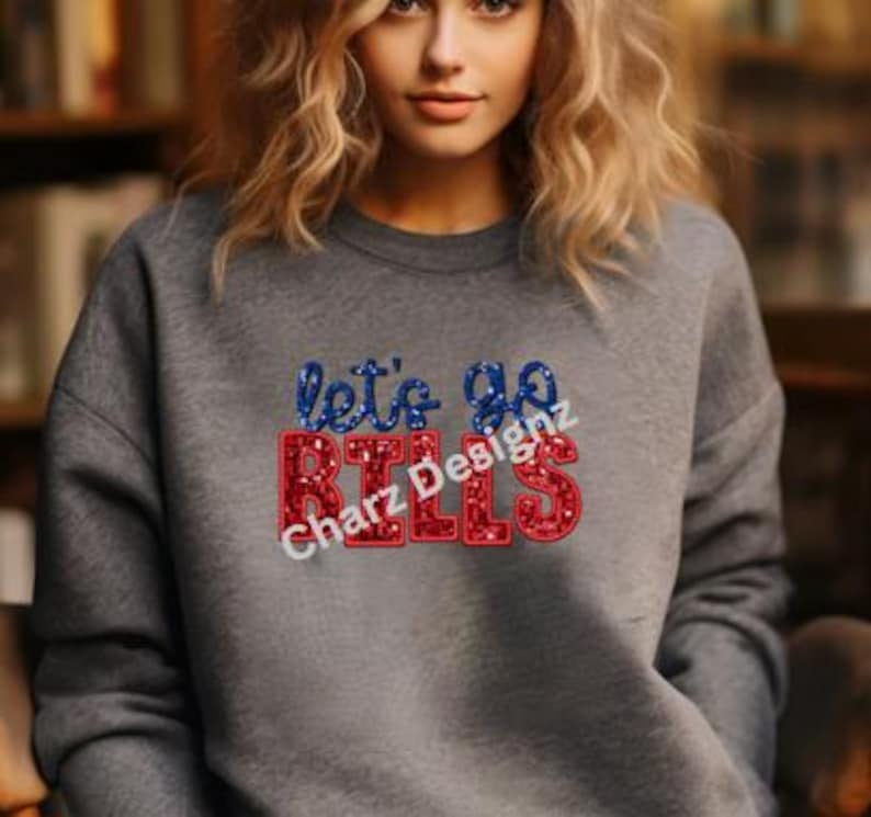 May include: A gray sweatshirt with the text "let's go BILLS" in red and blue glitter lettering. The sweatshirt also has the text "Charz Designz" in white. The sweatshirt is a crew neck style.