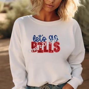 May include: White sweatshirt with the text "let's go Bills" in blue and red glitter lettering. The sweatshirt has a crew neck and long sleeves. The text is centered on the front of the shirt.