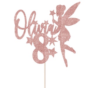 May include: Rose gold glitter cake topper featuring the name Olivia and the number 8, a fairy silhouette with wings and a wand, and stars.