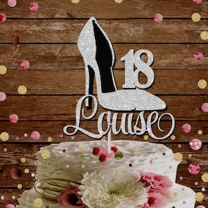 May include: Silver glitter high heel shoe cake topper with the number 18 and the name Louise written in cursive. The topper is on a white cake with pink and gold sprinkles.