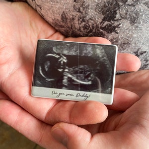 May include: A rectangular white magnet featuring a black and white ultrasound image of a fetus. Below the ultrasound, a light grey horizontal band contains the black printed text "See you soon Daddy!" in a cursive script font style.