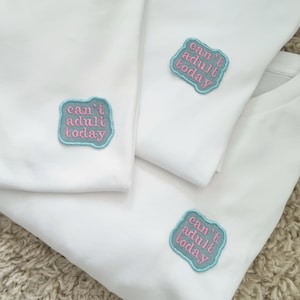 Könnte beinhalten: Drei weiße T-Shirts mit einem blauen und rosa gestickten Aufnäher mit der Aufschrift "can't adult today".
