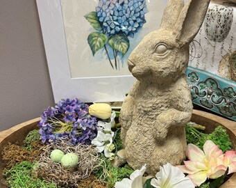 Peut inclure: Une statue de lapin beige est placée à côté d'un bol en bois rempli de mousse, de fleurs et d'un nid avec deux œufs verts. Une aquarelle encadrée d'hortensias bleus est en arrière-plan.