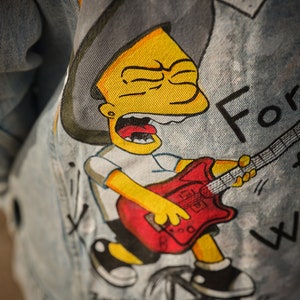 Può includere: Una giacca di jeans con un disegno dipinto a mano di un personaggio dei cartoni animati giallo che indossa un cappello da cowboy e suona una chitarra elettrica rossa. Il testo "For" è visibile sulla giacca.