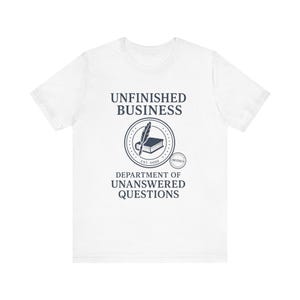 Puede incluir: Camiseta blanca con un gráfico azul marino. El gráfico incluye las palabras "UNFINISHED BUSINESS" sobre un diseño circular con una pluma, un libro y el texto "DEPARTMENT OF UNANSWERED QUESTIONS". El diseño también incluye la palabra "CONFIDENTIAL".