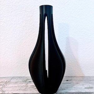 Peut inclure: Un vase décoratif noir avec un design ouvert unique. Le vase a un col fin et un corps évasé en forme de pétale. Il repose sur une surface en bois contre un mur blanc.