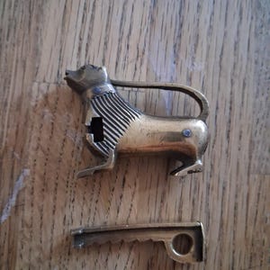 May include: A brass padlock shaped like a standing lion with a textured mane and a separate, matching brass key. The lion figure features a curved shackle extending from its back to its hindquarters, with a keyhole located on the chest area.