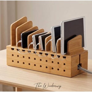 May include: A bamboo charging station with multiple slots for phones and tablets. The station has a rectangular base with several small holes and a series of angled dividers. White charging cables are connected to the devices. The Woodeenry logo is at the bottom.