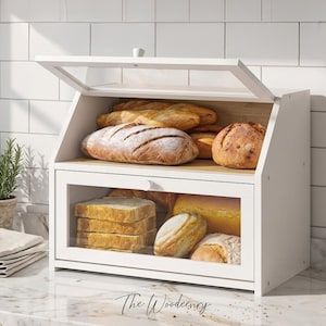 Puede incluir: Una panera de madera blanca con puerta y tapa de cristal transparente, llena de varios tipos de pan. La panera tiene un diseño sencillo y está sobre una encimera de mármol. El pan incluye hogazas, baguettes y pan rebanado.